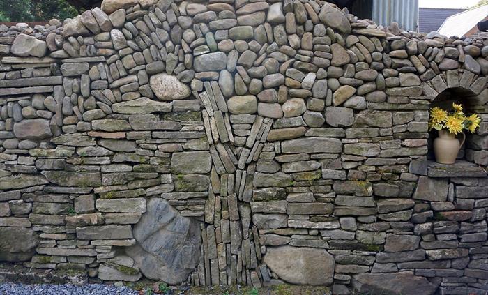 Handcrafted dry stone wall with intricate patterns and a small alcove holding a vase of yellow flowers.